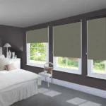 Two gray blackout roller blinds lowered over a large window overlooking a patio