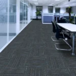 Dark gray and black modular carpet tiles with faint linear patterns laid in a hallway